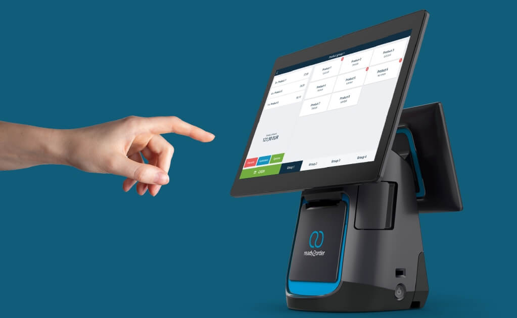 Eine Hand zeigt auf ein schwarzes Touchscreen-Pos-Terminal mit blauem Akzent und dem "ready2order"-Logo, das eine Verkaufsoberfläche anzeigt