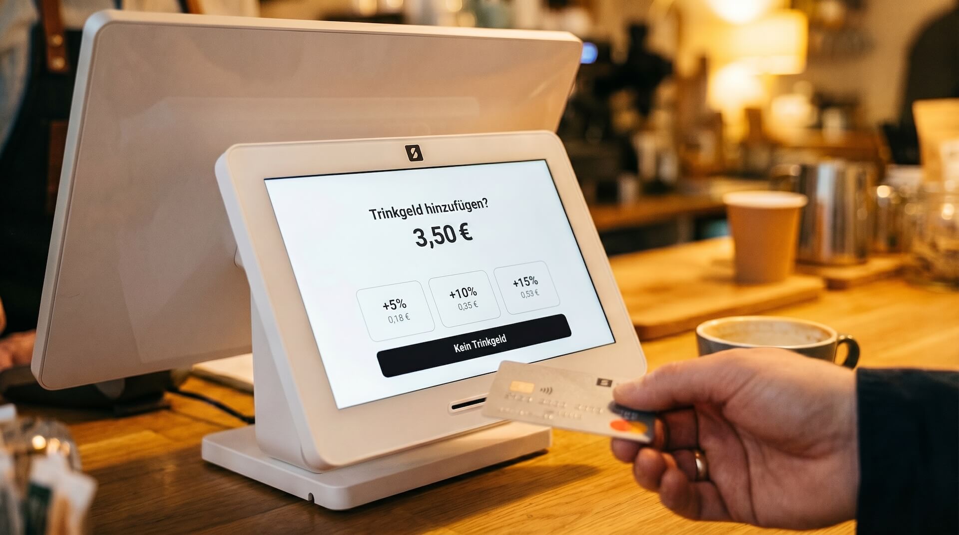 SumUp Kassensystem mit Doppeldisplay auf einer Café-Theke: Das Kundendisplay zeigt die Trinkgeld-Abfrage mit Optionen für 5, 10 und 15 Prozent, während eine Hand die Karte kontaktlos vor das Terminal hält.